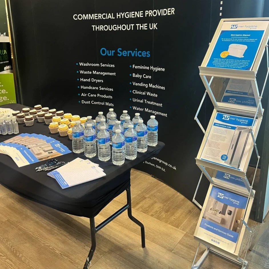 A black table at an event covered in cupcakes, bottled water, and brochures in front of a banner listing NWR Hygiene's services.