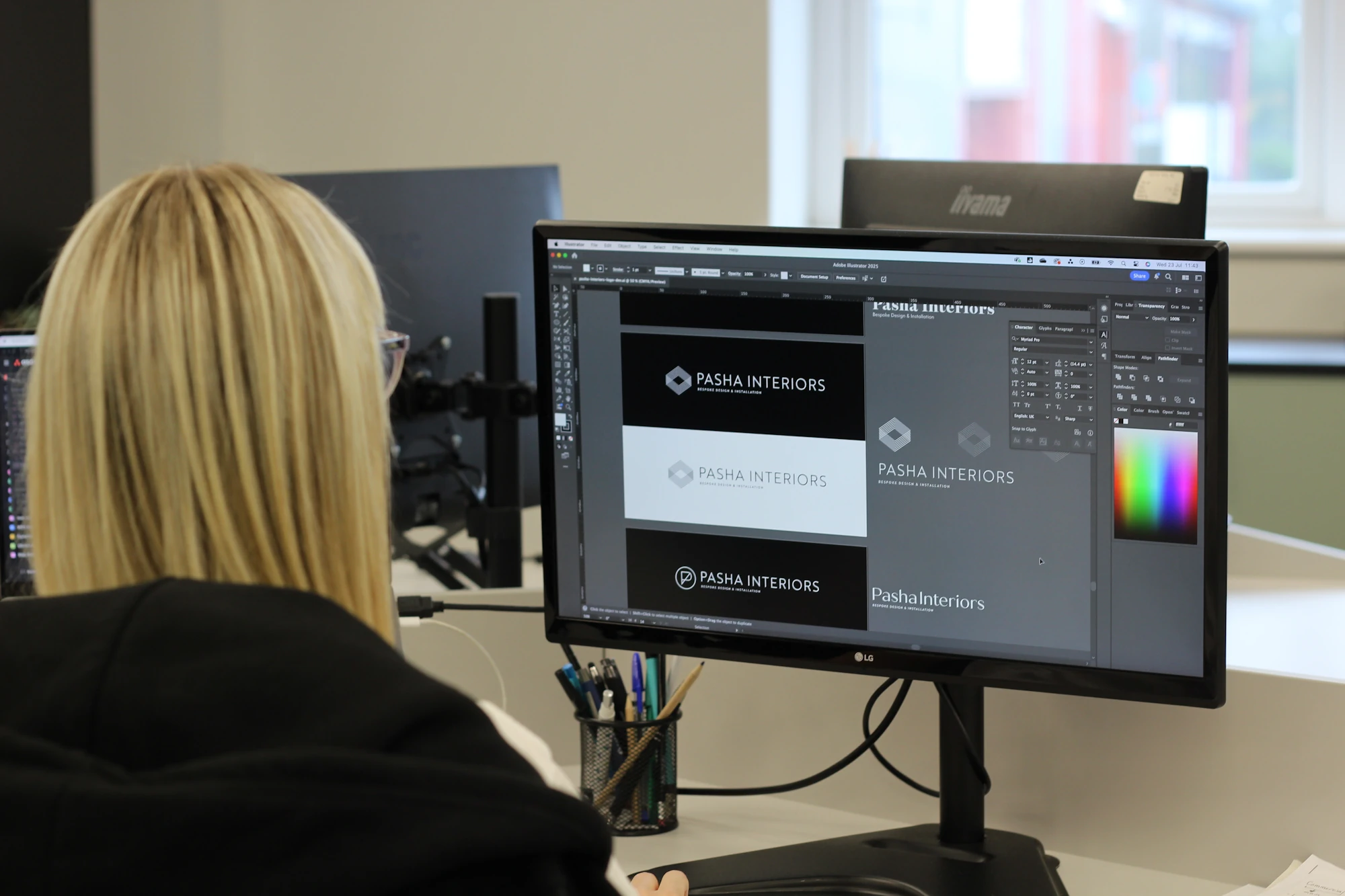 Shelley, Graphic Designer at TVG, working on a branding project for 'Pasha Interiors' on a large monitor. The screen shows various logo concepts and a colour palette in Adobe Illustrator.