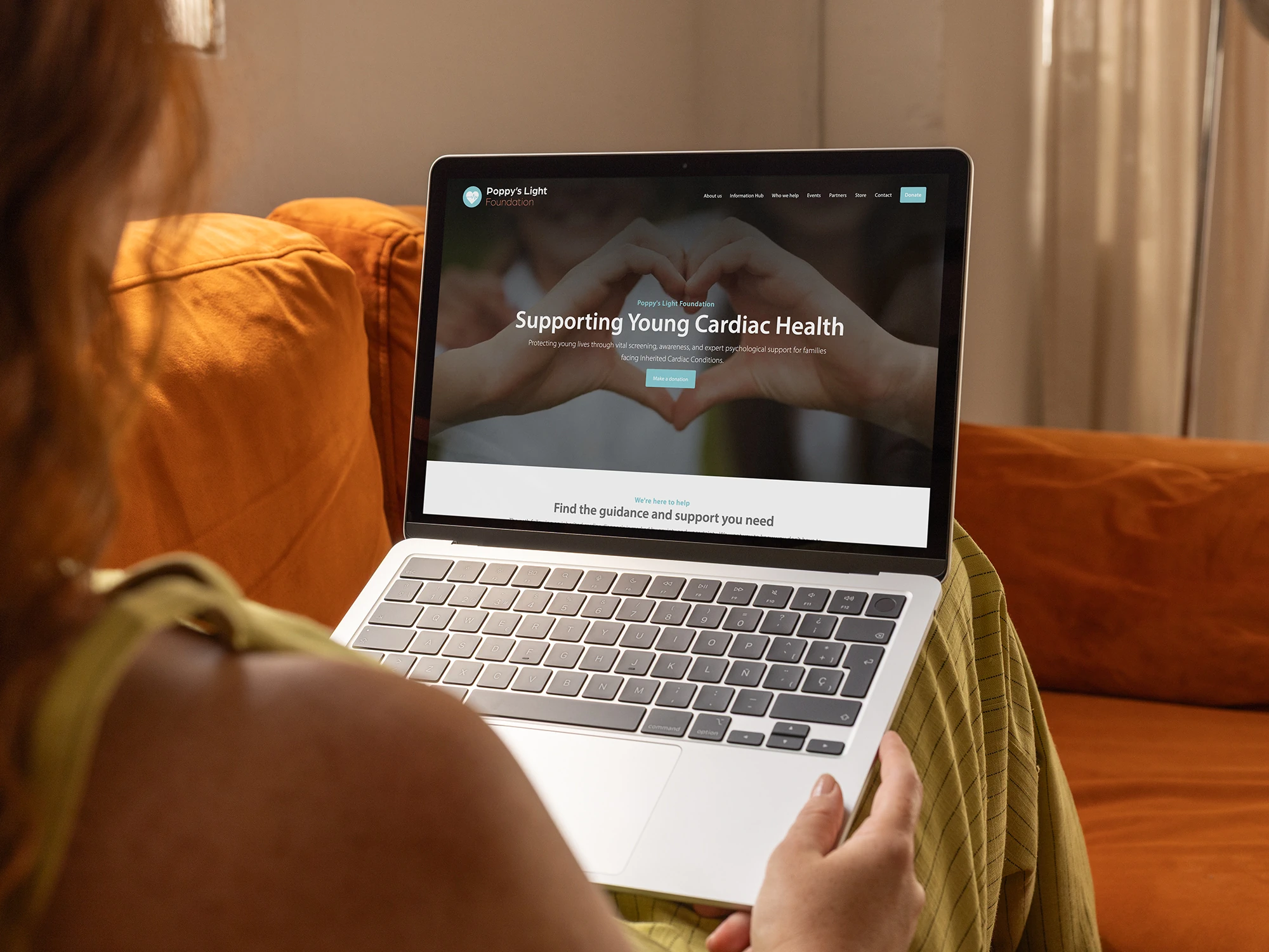 Woman sat on orange velvet sofa, looking at Poppy's Light Foundation website on laptop.