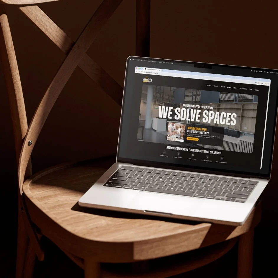 A laptop sitting on a wooden chair showing the Ambic Manufacturing home page and slogan.